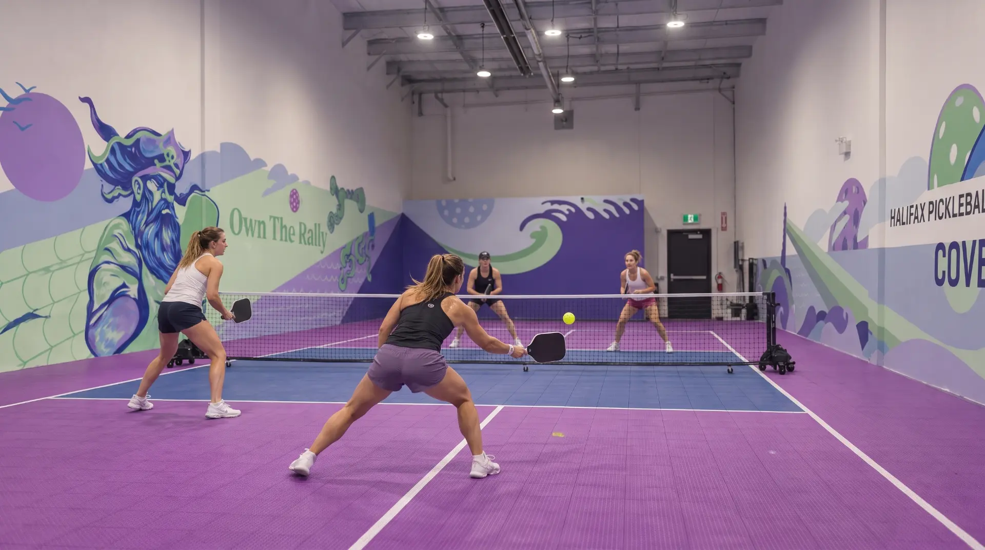 Women's doubles rally on the purple indoor court in front of the Own The Rally mural at Pickleball Cove