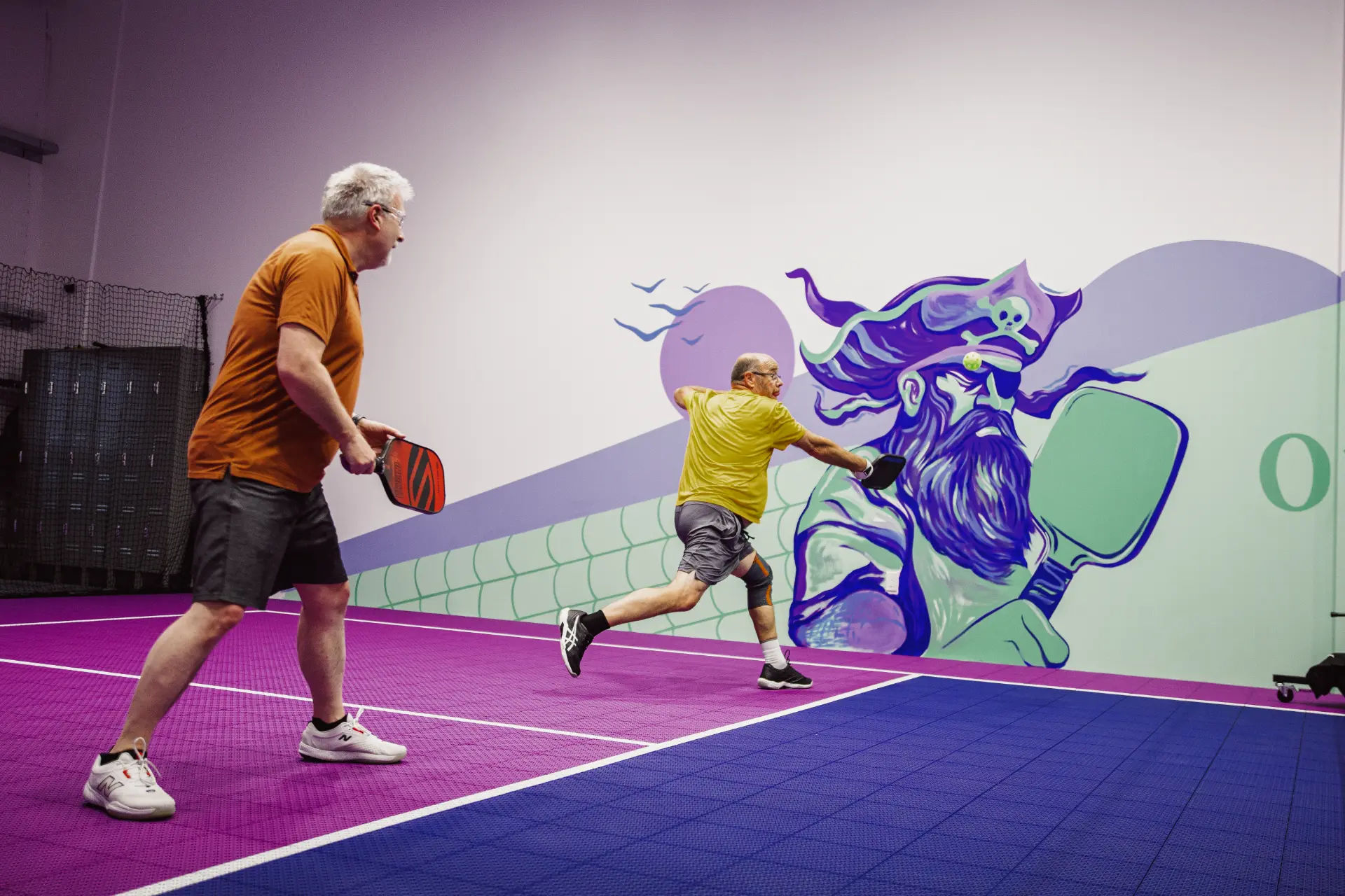 Two players mid-rally on the purple indoor court at Pickleball Cove, with the Roger the Pirate mural behind them